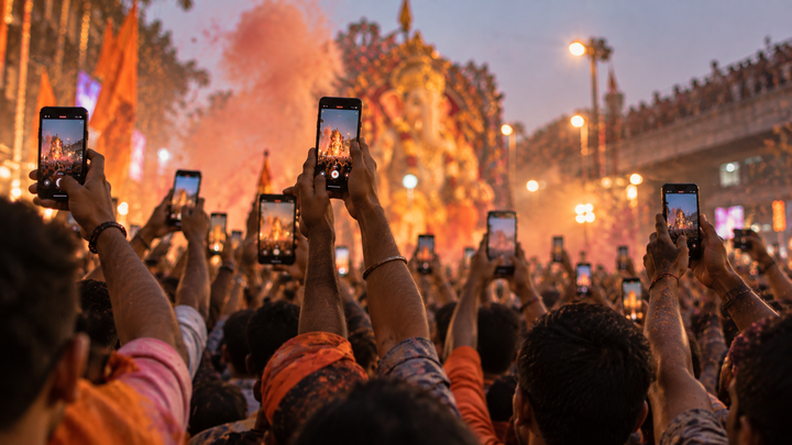 Raised hands holding phones above a Ganesh visarjan procession at dusk, gulal haze in the air